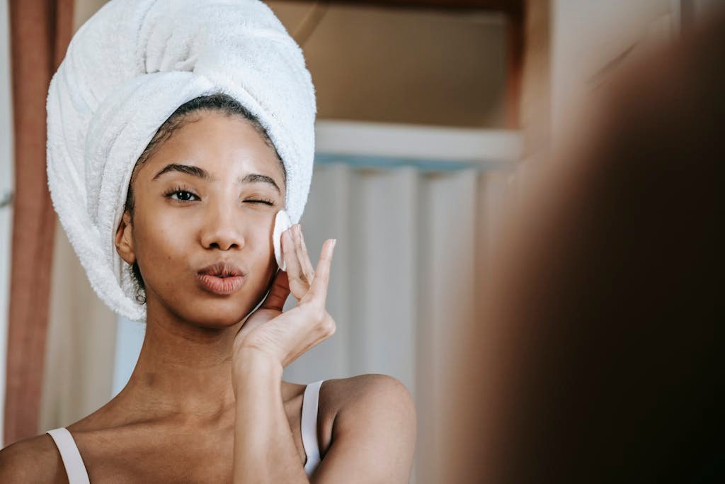 Charming ethnic female with folded lips winking while cleaning face with cotton pad during daily beauty routine