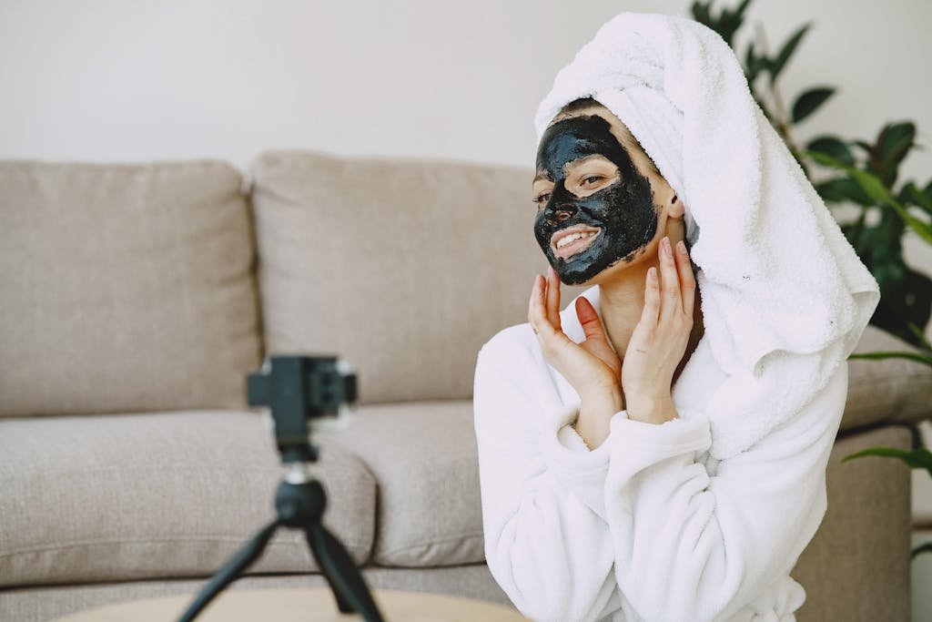 Positive female in white bathrobe and towel with black facial mask sitting near sofa in living room and taking photo on camera